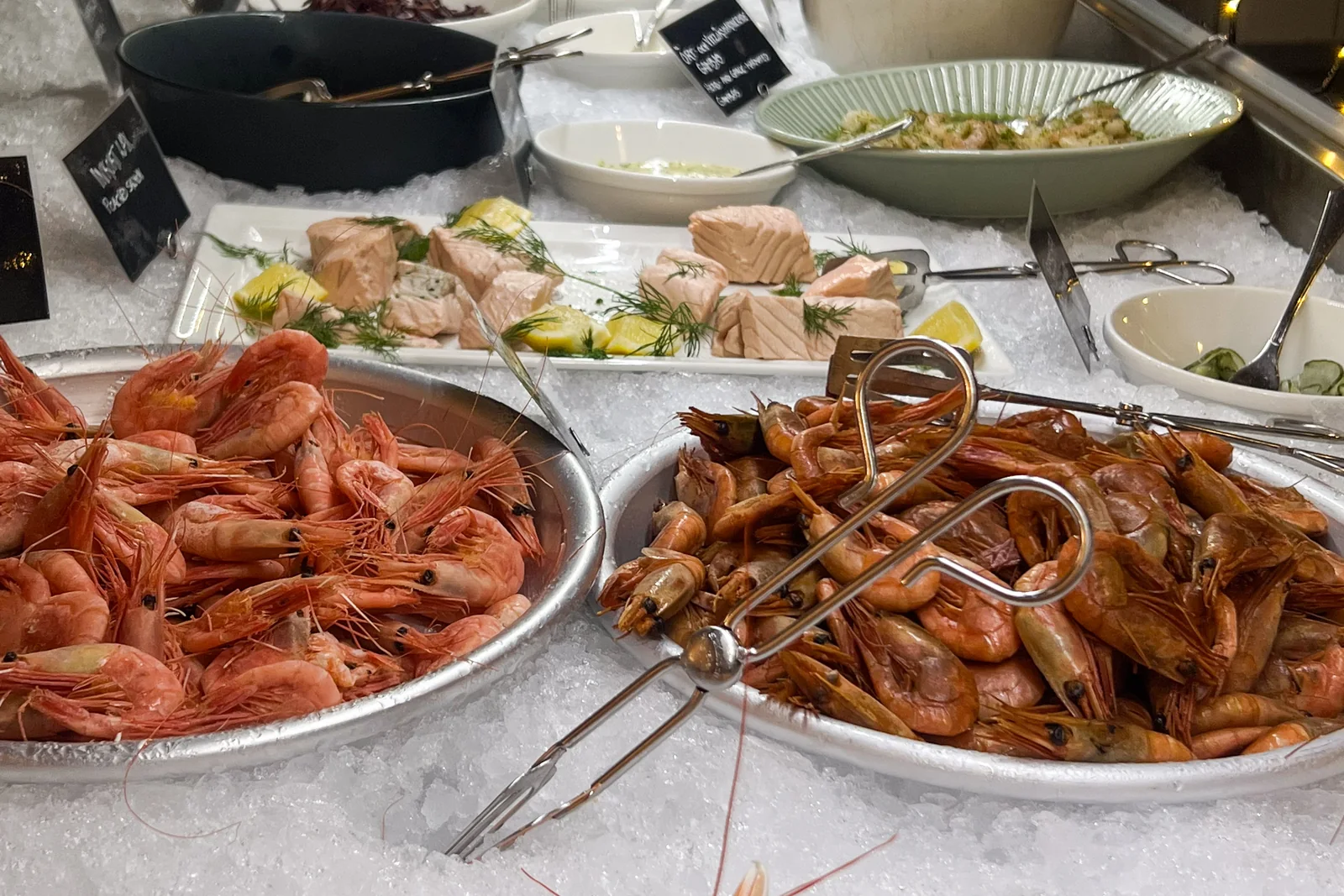 Two platters of prawns on ice, salmon with dill and lemon behind, chalk signs beside them