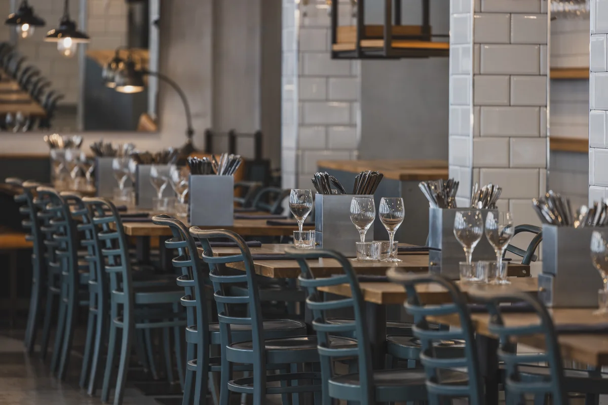 Long tables set for parties, blue-grey bentwood chairs against white tile and hanging lamps