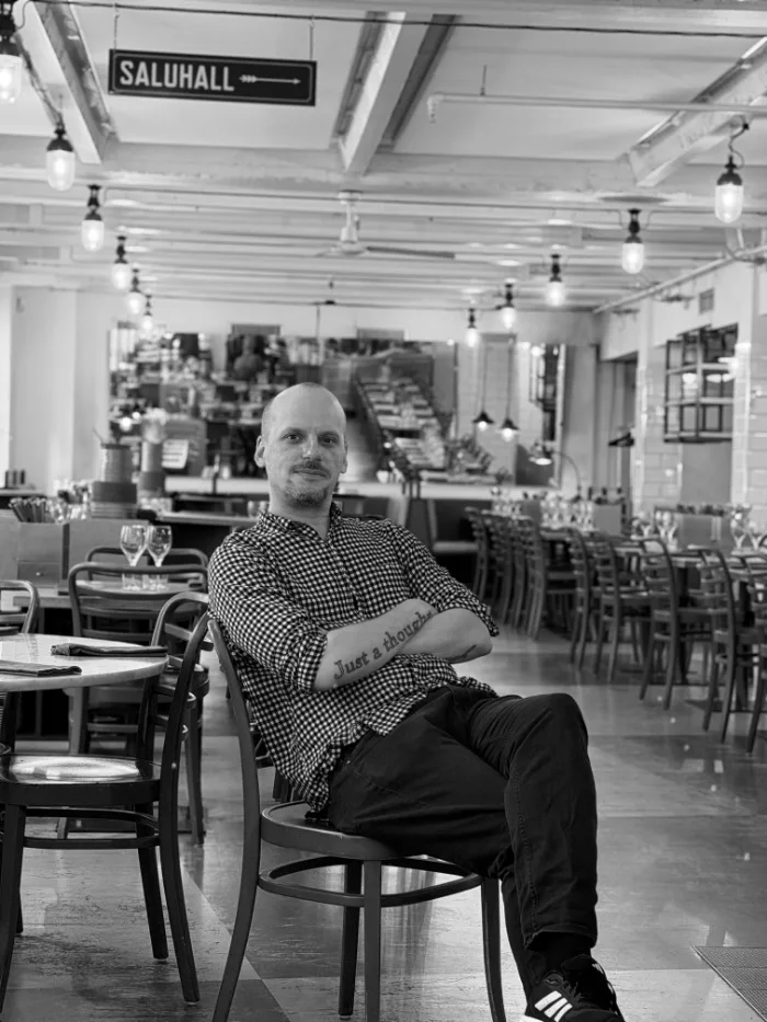A member of the B.A.R. team in a checked shirt, arms folded, a tattoo on the forearm, seated in the dining room under the Saluhall sign