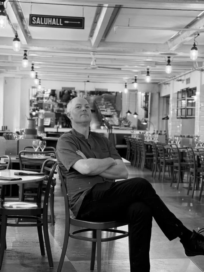 A member of the B.A.R. team in a dark shirt, arms folded, seated in the dining room under the Saluhall sign