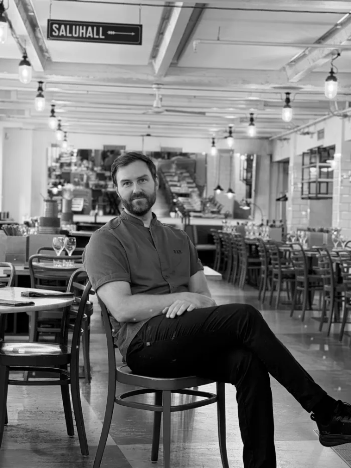 A member of the B.A.R. team in a dark polo shirt and a beard, seated in the dining room under the Saluhall sign