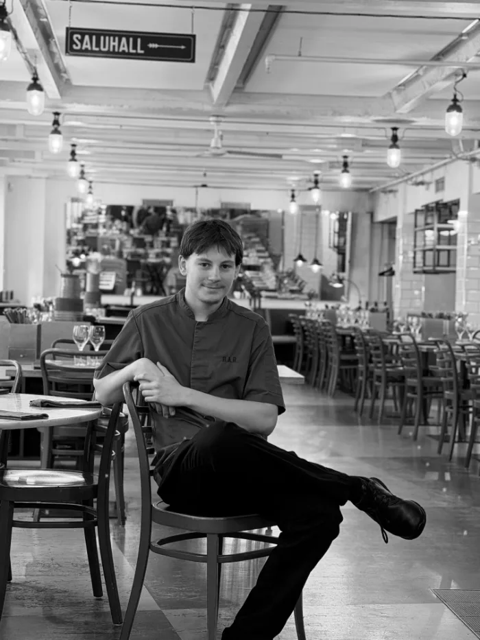 A member of the B.A.R. team in a dark chef's jacket, leaning on the back of the chair, seated in the dining room under the Saluhall sign