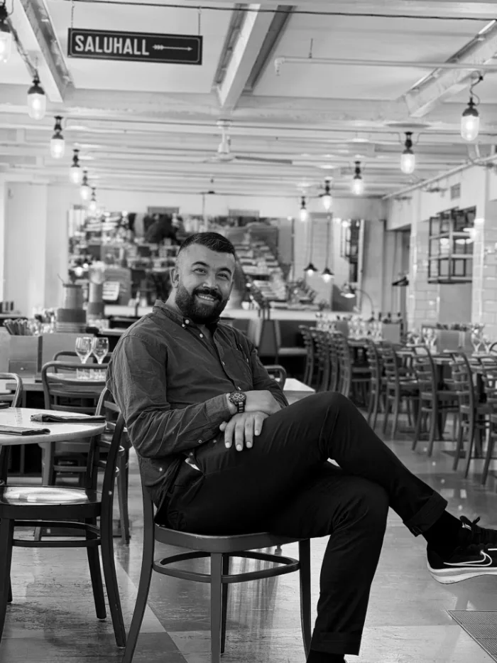 A member of the B.A.R. team in a dark shirt and a beard, leaning back, seated in the dining room under the Saluhall sign