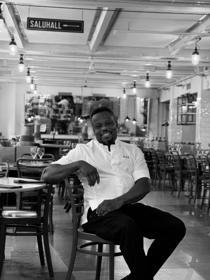 A member of the B.A.R. team in a white chef's jacket with B.A.R. embroidered on the chest, seated in the dining room under the Saluhall sign