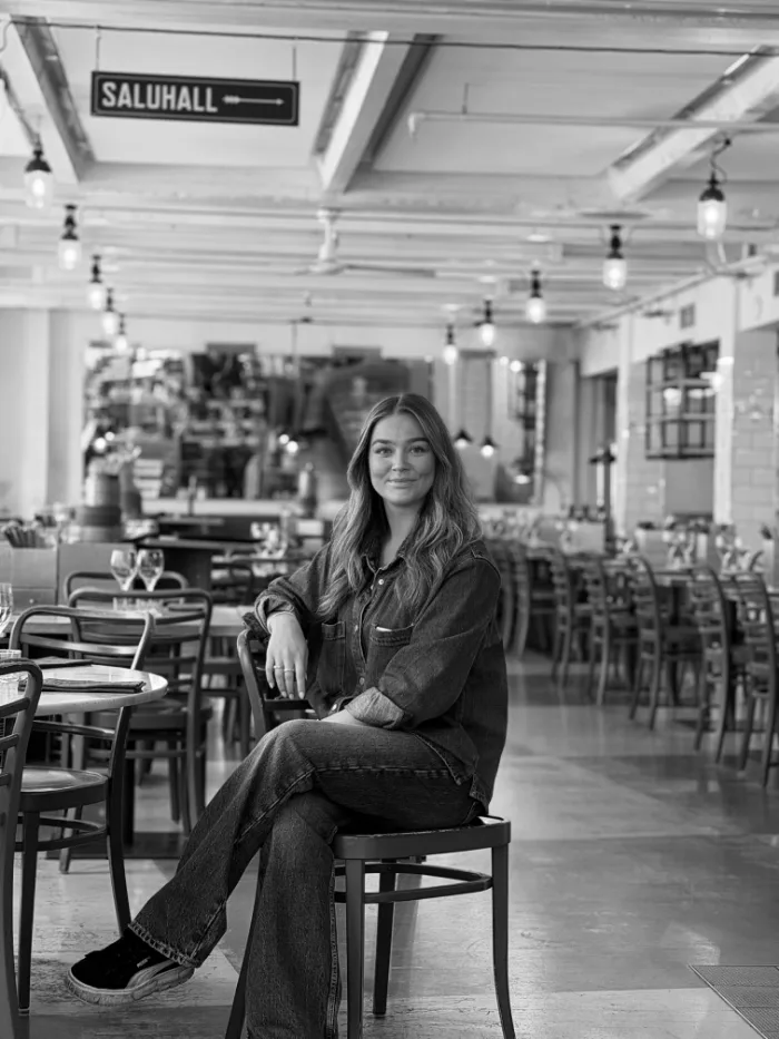 A member of the B.A.R. team in a light denim shirt and long hair, seated in the dining room under the Saluhall sign