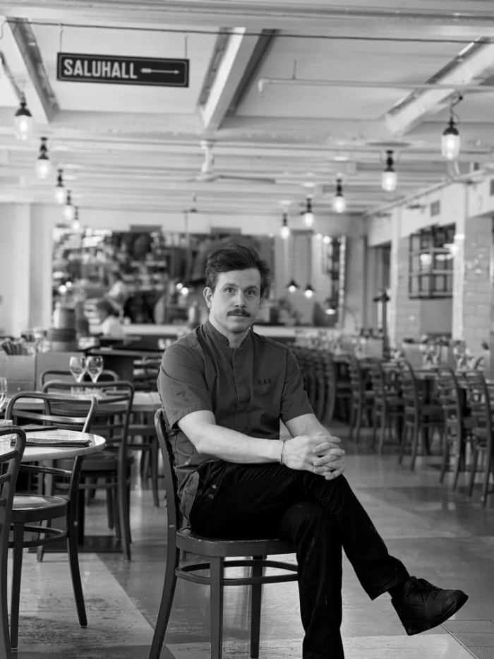 A member of the B.A.R. team in a dark shirt and a moustache, hands in their lap, seated in the dining room under the Saluhall sign