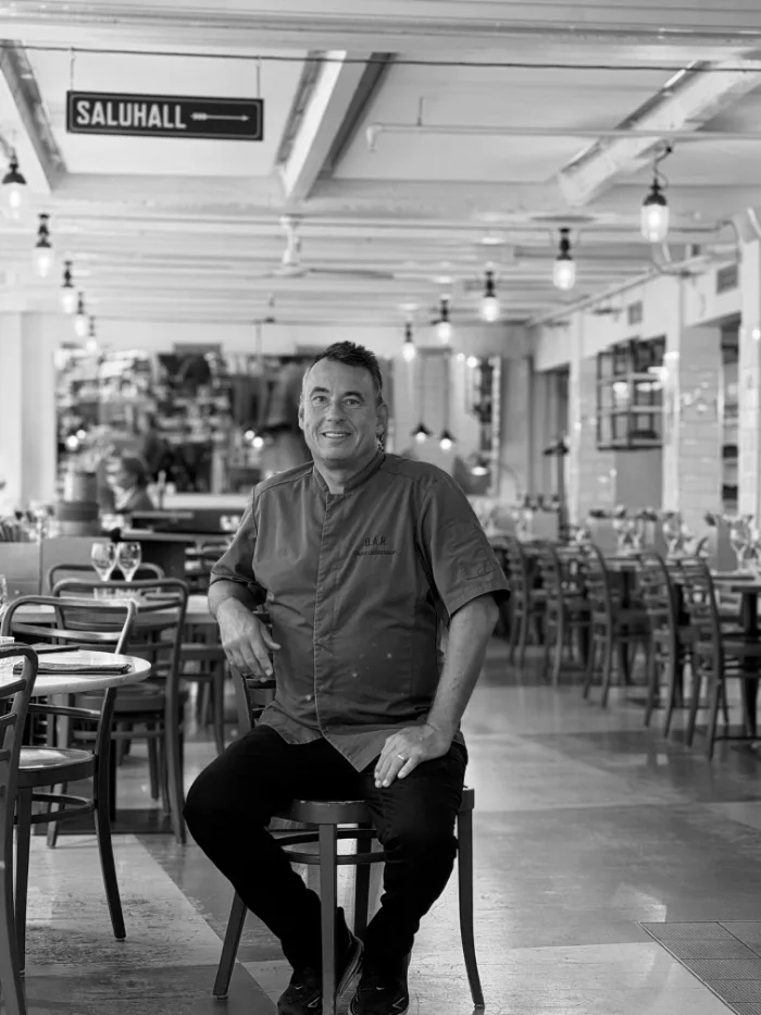 A member of the B.A.R. team in a dark chef's jacket, hands behind their back, seated in the dining room under the Saluhall sign