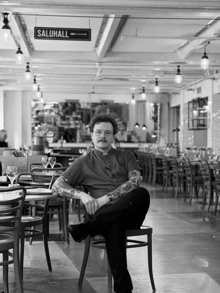 A member of the B.A.R. team in a dark shirt and a tattooed forearm, seated in the dining room under the Saluhall sign